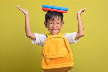Back to school. Cheerful Asian boy with backpack and carrying books on his head isolated on yellow...