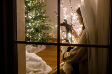 Merry Christmas! Beautiful woman in window hugging cute cat and looking at stylish gifts on...