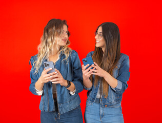 Studio shot portrait of young two caucasian women standing over red color background using mobile phone looking each other excited face expression. People lifestyle concept. Copy space.