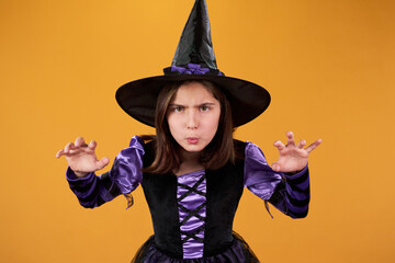 Cute little girl in witch dress and hat looking at camera and growling while casting spell against orange background