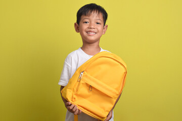 A happy Asian boy holding a school backpack isolated on yellow background. A cute kid preparing to study. Back to School concept