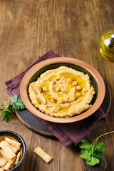 Hummus with sweet potato garnished with seasonings, olive oil and cooked chickpea grains in a brown ceramic plate on a brown wooden background. Served with crackers.