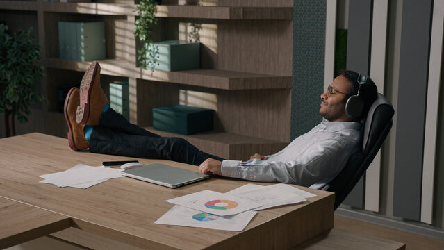 Calm relaxed American businessman Indian man Arabian worker entrepreneur in headphones listen music song in sound app legs on table office enjoy audio relaxing resting work break finish tranquil relax