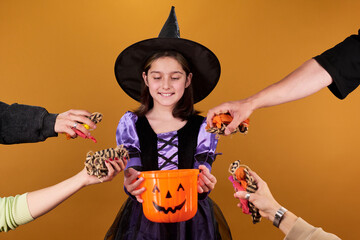 Smiling youthful girl in costume of Halloween witch looking at orange pumpkin shaped bucket while...