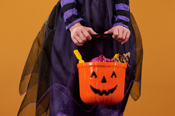 Hands of youthful girl in Halloween witch dress holding orange bucket with candies and other treats...