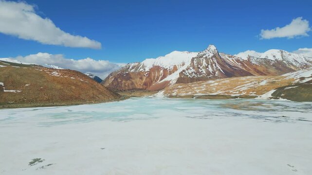 Aerial view of Suraj Tal Lake, partially frozen and surrounded by rugged, snow-capped peaks, with a winding path cutting through the stunning high-altitude terrain.
