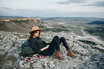 adventurous woman sitting on mountain ledge with backpack and hat, enjoying breathtaking view