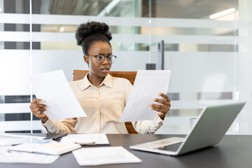 African American businesswoman at office desk examines two reports with concerned expression. She...
