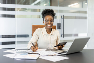Fototapeta premium Confident African American businesswoman with calculator, phone, laptop, financial documents in office. Shows professionalism, finance expertise, productivity. Ideal for business, finance themes.