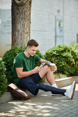 Fototapeta premium A young man with a prosthetic leg sits on the ground, sipping coffee and checking his phone in a sunny location.