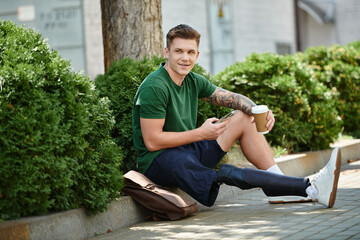 A young man with a prosthetic leg relaxes on a sunny day, enjoying coffee among lush greenery.