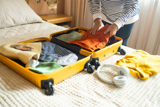 Woman packing suitcase on bed for a new journey packing list for travel planning preparing vacation. Open yellow suitcase with different family clothes and accessories on sofa.  - Powered by Adobe