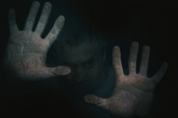 Horror scene of a man with hand against wet glass on dark background.