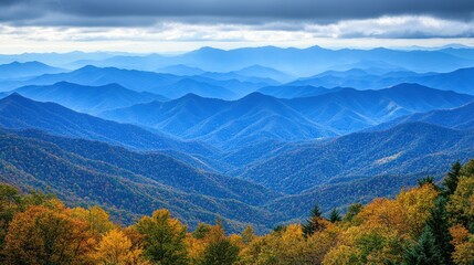 A panoramic view of a mountain range with fall foliage in the foreground and a cloudy sky.