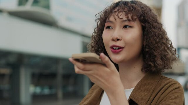 Happy Asian Woman With Curly Hair Recording Voice Message On Her Phone While Standing In Center. Young Woman With Nose Piercing Speaking Into Speaker Against Background. People and Technology Concept
