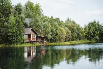Obraz premium Nestled beside a serene lake, a quaint small wooden house stands gracefully, framed by lush green trees, creating a stunning view of the breathtaking natural landscape all around