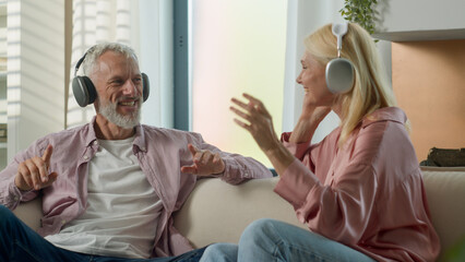 Joyful senior elderly Caucasian married couple grandparents man woman listening music using headphones dancing fooling around having fun carefree grandmother grandfather enjoying weekend at home couch
