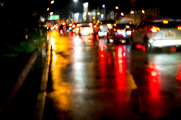 A blurry image of a busy street at night with cars and a few pedestrians. The image has a moody and somewhat chaotic feel to it, as the cars are moving quickly and the rain is making the street slick