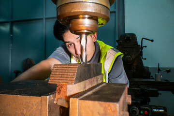 An industrial worker operating a drill press in a workshop. The focus is on the precision and concentration required to handle machinery. The worker wears a safety vest, ensuring a secure work.