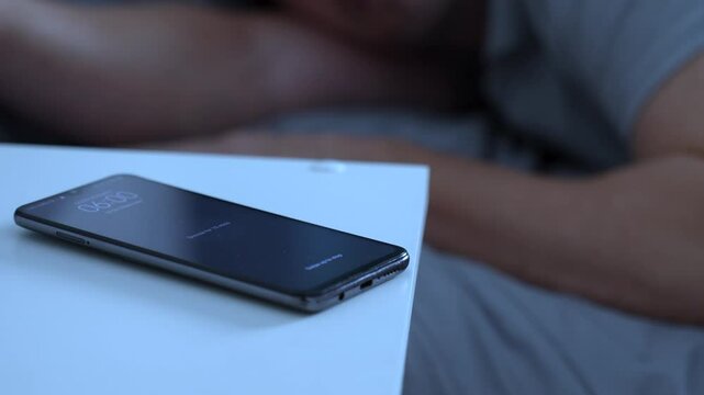 Man waking up, turning off alarm clock to snooze mode on smartphone screen. Sleepy man touching phone with alarm at 6 o'clock morning