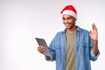 Smiling african young teenage boy man waving with a hand saying hello while having videocall conversation online on digital tablet wearing Santa`s red hat for Christmas New Year isolated on white