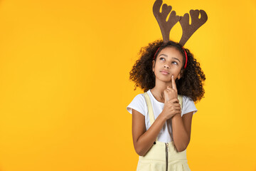 Photo of pretty little girl kid with head horn looking doubtful isolated on yellow background. Deer costume for Christmas New Year celebration. Dreaming about presents