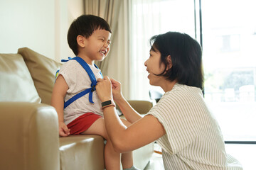 Obraz premium Asian Mother adjusts her young son backpack as he sits on the couch in a living room.