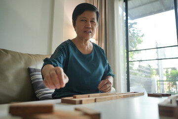 Asian Senior woman concentrating while arranging wooden blocks on a white table in a living room.