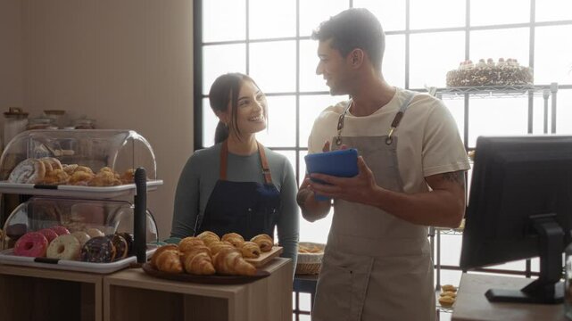 Man and woman in a bakery looking at a tablet while preparing pastries and donuts