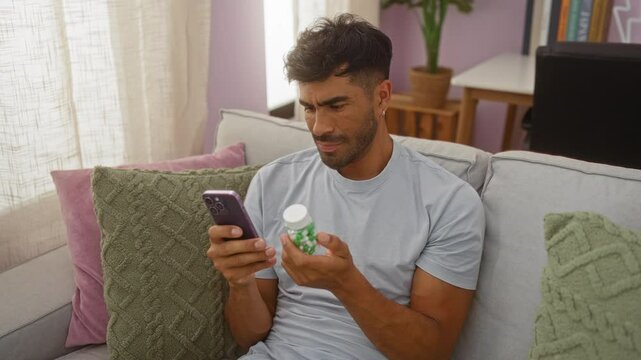 Young man indoors looking at a phone while holding a bottle, sitting on a couch in a living room with pillows and furniture in the background, appearing to be considering or reading something.