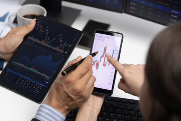 Cropped with focusing on phone with hand pointing with discussing dynamic stock market in two business traders on screens on real time currency rate investment risk data at modern office. Postulate.