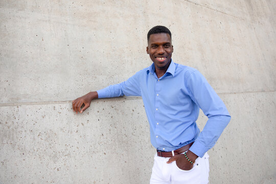 Portrait Of Handsome Smiling With Teeth African American Man wearing White Jeans and Blue Shirt, Brown Belt Outdoor, Concrete Wall on Background. Copy Space Fashionable Male, Ivorian. Horizontal
