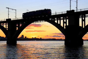 Train, diesel locomotive goes on the arched railway bridge on the river at a beautiful sunset, silhouette, the city Dnepr, Dnipro, Ukraine.