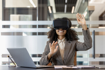 Young African American woman sitting in the office at the desk in a suit wearing virtual glasses and gesturing with her hands