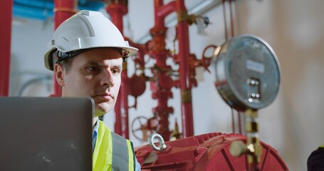 Engineers Analyzing Equipment inspects industrial piping while using a laptop in a Factory, Focused...