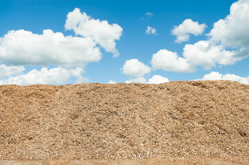 The photo shows a mountain of wood chips close-up against a blue sky and clouds