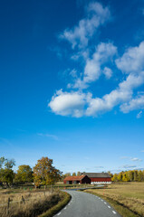 Naklejka premium Clouds in blue sky over Sweden countryside in autumn season, Narrow asphalt vinding country road among dry fields leading to wooden red farmhouses