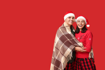 Happy young couple in Christmas pajamas and Santa hats wrapped in plaid on red background