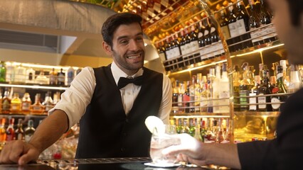 Smart bartender serving drink gin fizz with lemon sliced to VIP guest while waiting friend at luxurious counter bar. Barman providing excellent service at vibrant nightlife party atmosphere. Vinosity.