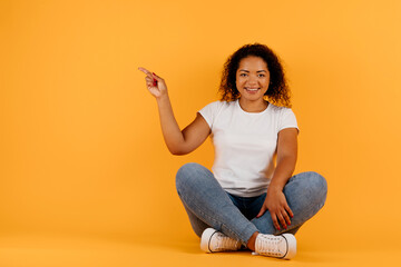 Nice Promo. Happy Black Woman Pointing At Copy Space Over Yellow Background, Sitting On Floor In Studio, Panorama. High quality photo