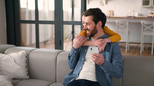 Cheerful Caucasian daughter little child surprise hug embracing distracting dad parent male father man type mobile phone kid smile naughty girl require attention fooling around family laughing at home