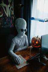 Teenager in Halloween full-body freaky white costume with carved pumpkin laptop on table studying online