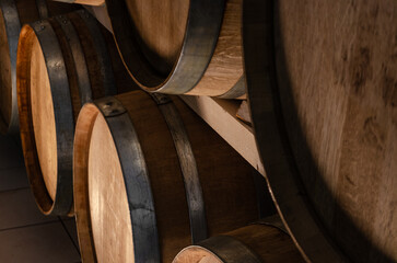 A close-up of several wooden wine barrels stacked in a cellar. The barrels are made of dark brown wood with silver metal hoops. The image has a warm, rustic feel.