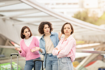 Three friends express different emotions while waiting at a train station, highlighting their contrasting opinions about travel plans amidst a stylish architectural backdrop.