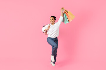 A young Asian woman joyfully holds colorful shopping bags while balancing on one leg. She wears a...