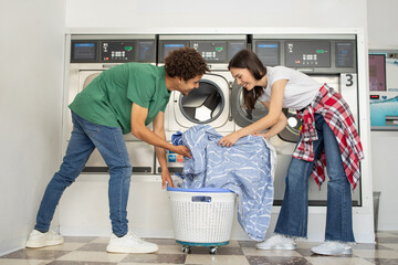 A young couple is enjoying their time at a laundromat, happily loading a washing machine with...