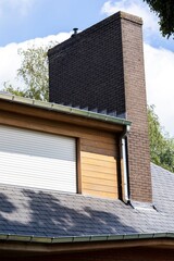 A residential building with a dark brick chimney, white roller shutter, wooden siding, and metal gutters. The roof is covered with gray shingles, with trees and a bright blue sky in the background.