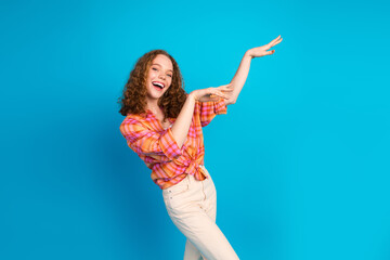 Naklejka premium Cheerful young woman with curly red hair gesturing and smiling in a bright outfit against a blue background.