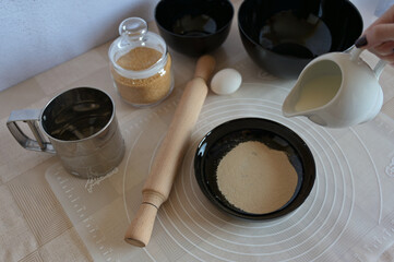 Baking ingredients for baking sweet buns, yeast in black bowl dishes, creamer with milk in a woman hand, rolling pin on a table