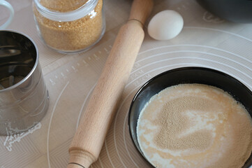 Close-up baking ingredients for baking sweet buns, yeast and milk in black bowl dishes, rolling pin on a table